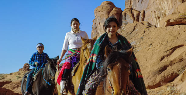 mehrere junge indianer (navajo) reiten pferde durch die monument valley wüste in arizona/utah neben einer großen felsformation an einem klaren, hellen tag - monument valley navajo mesa monument valley tribal park stock-fotos und bilder