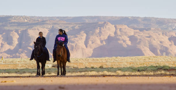 zwei indianerkinder reiten ihre pferde durch die wüste von arizona mit bergen/mesas hinter ihnen bei sonnenuntergang - monument valley navajo mesa monument valley tribal park stock-fotos und bilder