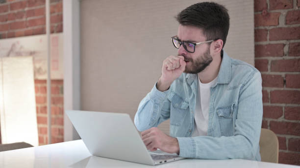 istock Sick Young Man in Glasses Working on Laptop and Coughing 1219309547