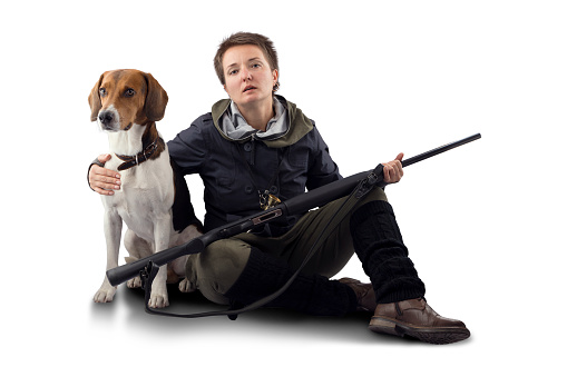 A woman with a short haircut, in a blue windbreaker, holds a gun in her hands, sits on a white background. A dog of the Estonian hound breed is sitting nearby.