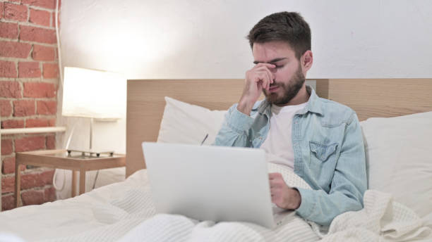 istock Young Man with Headache working on Laptop in Bed 1219309648