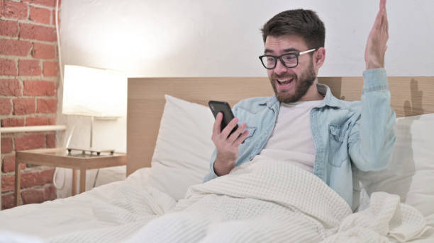 young man in glasses celebrating success on smartphone in bed - 15855 imagens e fotografias de stock