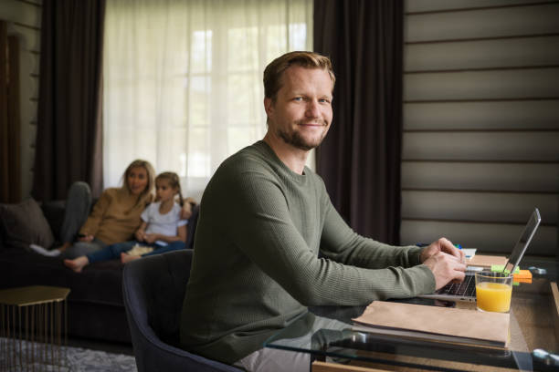 homem sorridente de meia idade olhando para a câmera enquanto trabalha no computador portátil sentado na mesa em casa. sua esposa e filha assistindo tv no sofá ao fundo - looking at camera smiling desk isolated - fotografias e filmes do acervo