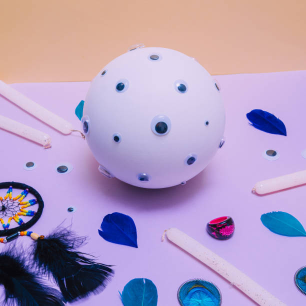 istock A white fortune telling ball with many eyes, a dream catcher, glittering candles, a ring and feathers on a light yellow and purple or lilac background. bstract minimal pop art. 1219325035