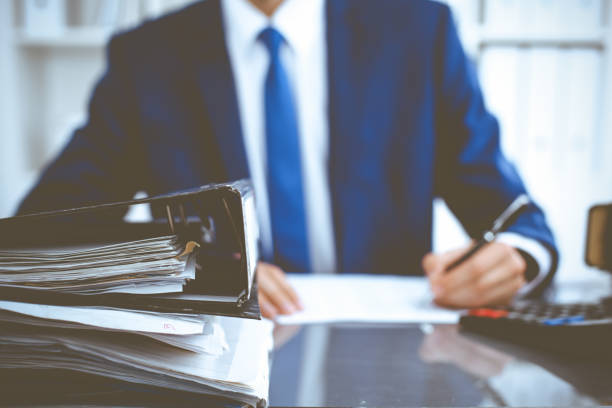 istock Binders with papers waiting to be processed with businessman or bookkeeper back in blur. Accounting planning budget. Audit, insurance and business concept 1219336947