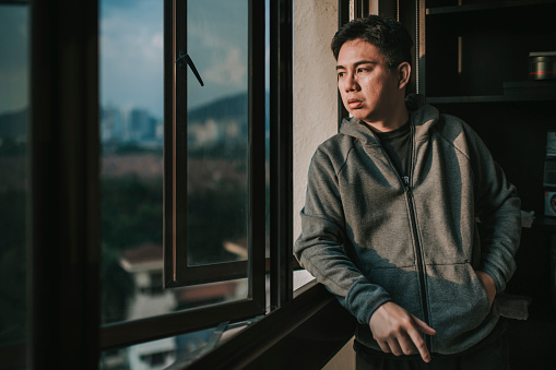 an asian chinese mid adult man looking outside of window from his home during sunset with serious facial expression with hooded shirt