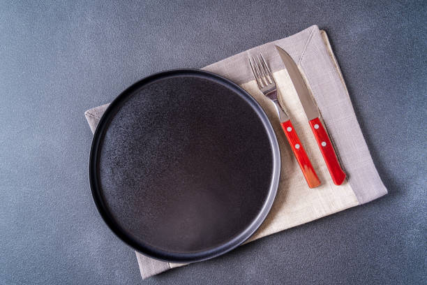 istock Empty black and cutlery plate over napkin on brown wooden table top view, copy space for text. 1219351707