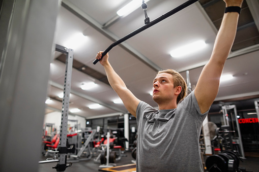 Young caucasian male exercising while in the gym. He is using a cable lateral pulldown machine to train his back.