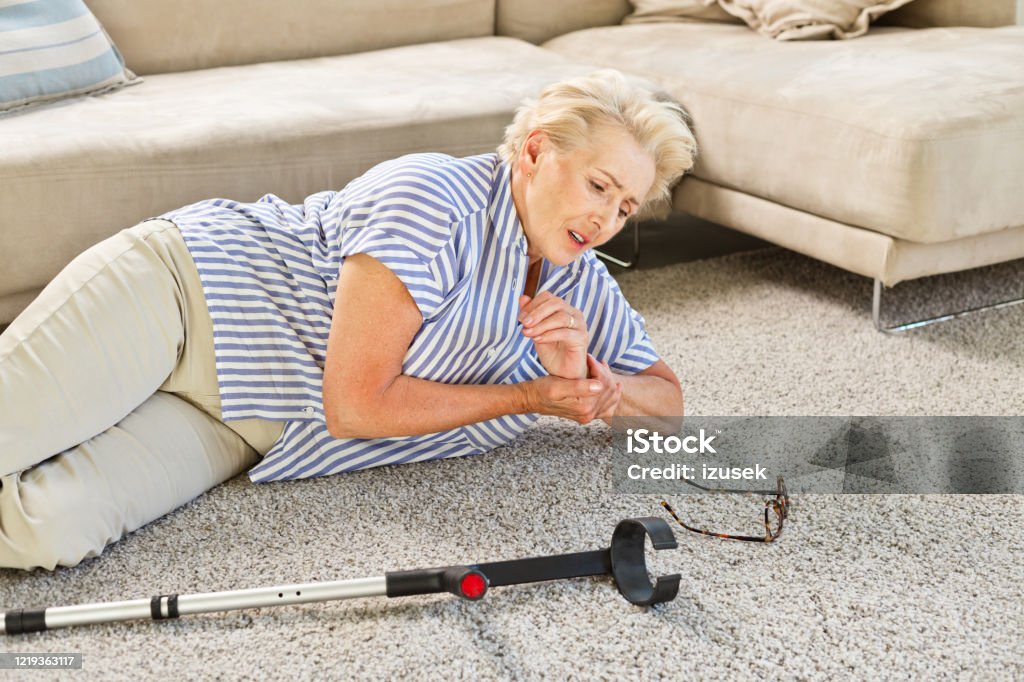 Elderly lady lying on the floor after moving down Tried and worried senior woman lying on the carpet in the living room and holding her wrist. Elderly lady falling down at home. Crutches on the floor. Senior Adult Stock Photo