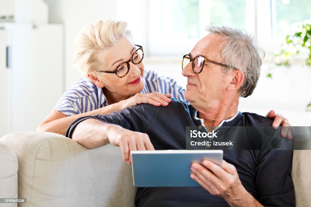 Friendly senior couple at home Elderly man sitting on sofa in the living room at home and holding digital tablet in hands, looking at his wife. Senior couple spending time together. Choosing Stock Photo