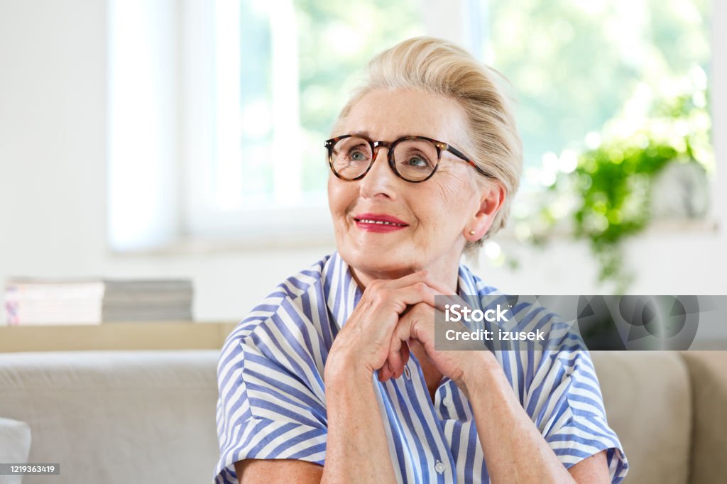 Portrait of beautiful elderly lady at home Attractive senior woman sitting on sofa in the living room at home. Elderly lady looking away and smiling. Active Seniors Stock Photo