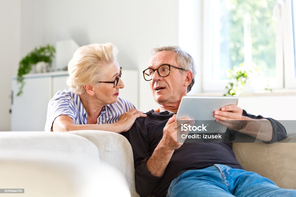 Senior couple spending time together at home Elderly man sitting on sofa in the living room at home and holding digital tablet in hands, talking with his wife. Senior woman looking at her husband. Home Interior Stock Photo