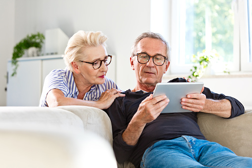 Elderly man sitting on sofa in the living room at home and showing something on digital tablet his wife. Senior woman peeking on screen.