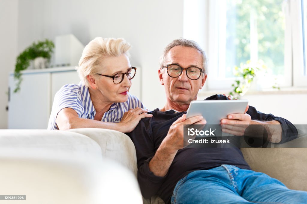 Senior couple watching digital tablet together at home Elderly man sitting on sofa in the living room at home and showing something on digital tablet his wife. Senior woman peeking on screen. Retirement Stock Photo