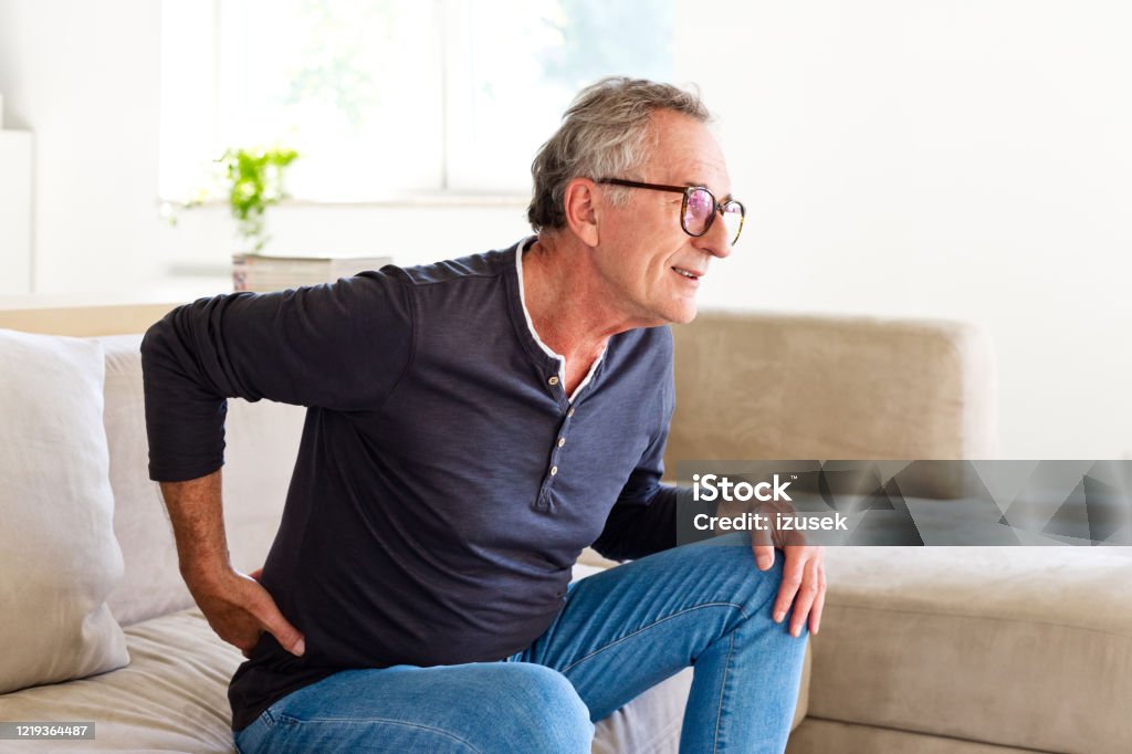 Senior man suffering backache Worried senior man sitting on sofa at home and touching his back. Elderly man suffering lumbar spine. Backache Stock Photo