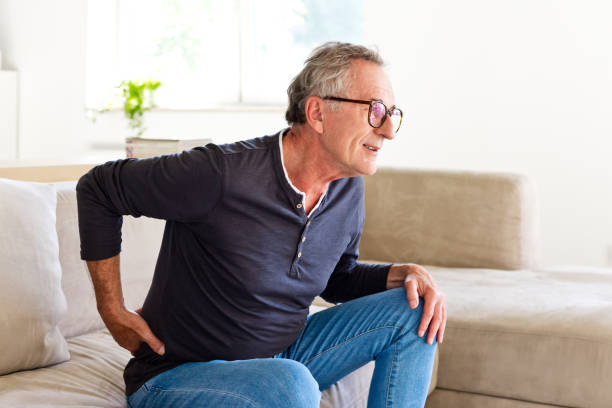Senior man suffering backache Worried senior man sitting on sofa at home and touching his back. Elderly man suffering lumbar spine. back pain stock pictures, royalty-free photos & images