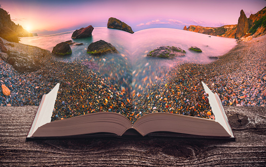 Sunrise over the sea bay with colorful pebble beach on the pages of an open magical book. Majestic nature. Travel concept.