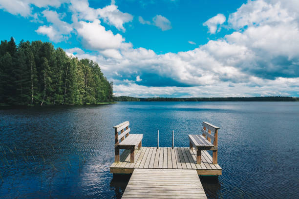 dos sillas de madera banco en un muelle de madera con vistas a un agua azul del lago con bosque verde y cielo nublado - finland lake summer couple fotografías e imágenes de stock