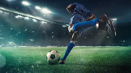 Professional football or soccer player in action on stadium with flashlights, kicking ball for winning goal, wide angle. Concept of sport, competition, motion, overcoming. Field presence effect.
