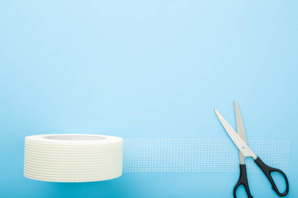 Scissors and white roll of mesh for split or seam on wall or ceiling on light blue table background. Pastel color. Closeup. Empty place for text. Top down view.