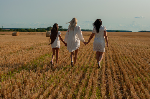 Mother and her two daughters in embroidered shirts walk along a wheat field, holding hands a view from the back