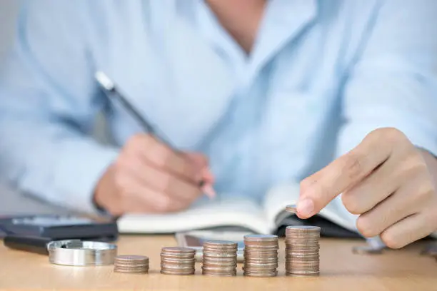 Photo of One hand putting coins stacking and one hand writing record for growth business and saving investment concept.