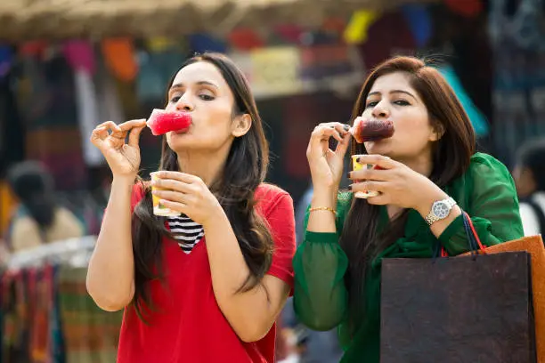 Two Indian women eating flavored ice gola dipped in syrup at street market