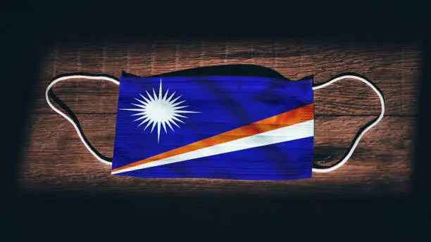 Marshall Islands National Flag at medical, surgical, protection mask on black wooden background. Coronavirus Covid"u201319, Prevent infection, illness or flu. State of Emergency, Lockdown...