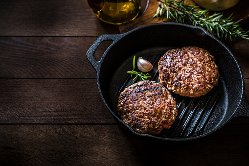 High angle view of a cooking pan with two grilled burger meats surrounded by a rosemary stick and an olive oil bottle on a rustic wooden table. Objects are at the right side of the image leaving a useful copy space at the left side. Low key DSLR photo taken with Canon EOS 6D Mark II and Canon EF 24-105 mm f/4L