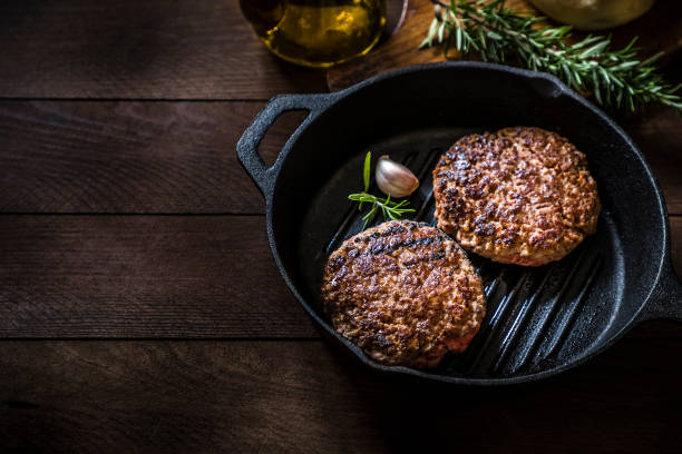 Grilled burger High angle view of a cooking pan with two grilled burger meats surrounded by a rosemary stick and an olive oil bottle on a rustic wooden table. Objects are at the right side of the image leaving a useful copy space at the left side. Low key DSLR photo taken with Canon EOS 6D Mark II and Canon EF 24-105 mm f/4L veggie burger stock pictures, royalty-free photos & images