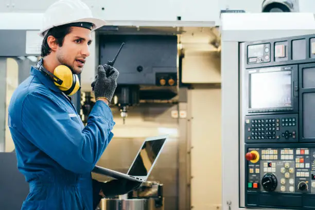 Photo of Male industrial engineer or technician in safety hard helmet and uniform using laptop checking on machine and radio phone calling team worker, heavy technology invention industry manufacturing factory