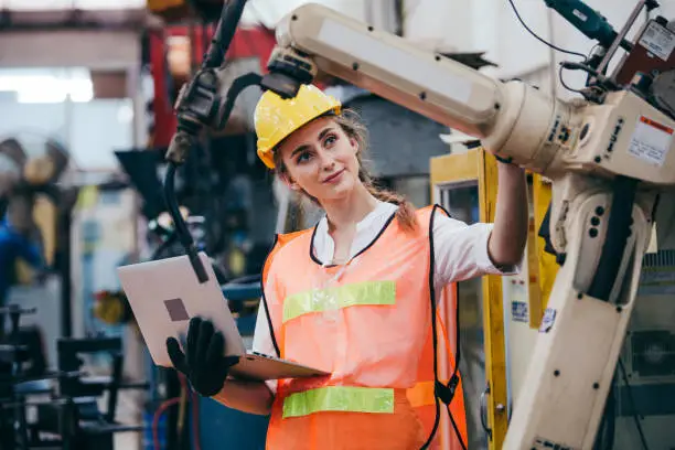 Photo of Female industrial engineer or technician worker in hard helmet and uniform using laptop checking on robotic arm machine. woman work hard in heavy technology invention industry manufacturing factory