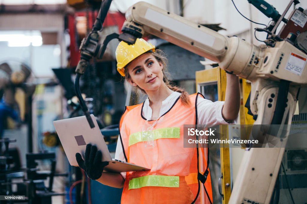 Kvinnlig industriingenjör eller teknikerarbetare i hård hjälm och uniform med hjälp av laptop kontroll på robotarm maskin. kvinna arbetar hårt i tung teknik uppfinning industrin tillverkningsfabrik - Royaltyfri Maskineri Bildbanksbilder