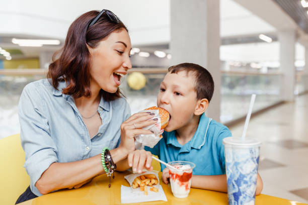 母と彼女の息子はモールで一緒にファーストフードランチを持っています - overweight child eating hamburger ストックフォトと画像