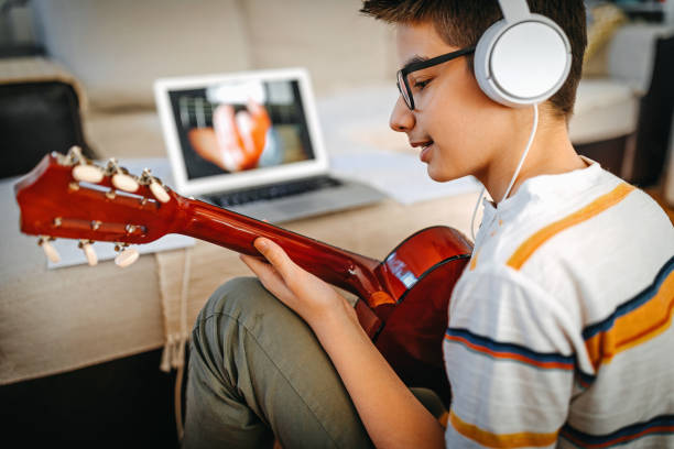 Teenage boy having online guitar lesson Teenage boy learning to play guitar watching online tutorial on laptop while listening to music on headphones. headphones plugged in photos stock pictures, royalty-free photos & images