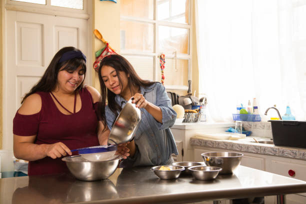 jeunes femmes hispaniques cuisinant - cuisson à la maison - soeurs heureuses cuisinant - activités à la maison - rolling dough pastry apron photos et images de collection