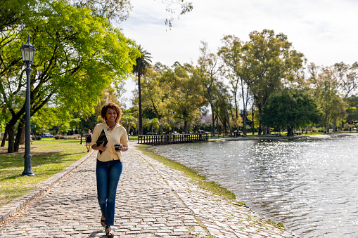 A woman is walking along the river in Buenos Aires. She is carrying a digital tablet and holding a reusable travel mug.