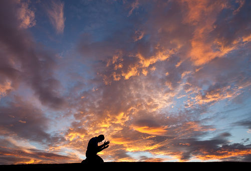 A silhouette of an unrecognizable man on his knees in prayer. Image taken in Nova Scotia, Canada. Side view. Man is in his 40s.