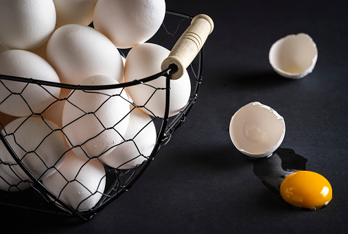 Chicken eggs in a metal basket on a black wooden table. Cracked egg directly on table with the yellow yolk and broken eggshell. Concept image for the proverb: don't put all your eggs in one basket