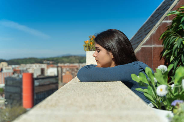 Woman looking at the street from her balcony during lockdown for coronavirus. She is alone, pensive and leaning on the railing.