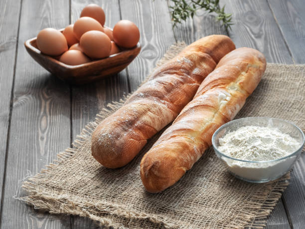 Two fresh french baguettes made from white flour next to chicken eggs and flour, baking ingredients. Bread located on burlap, rustic dark wooden background. Close-up. Horizontal layout