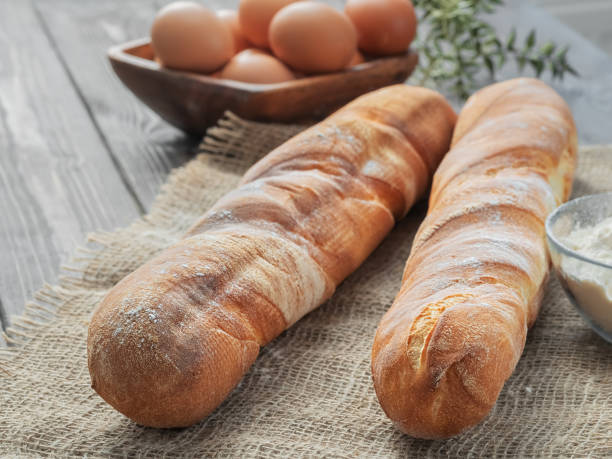 Two fresh french baguettes made from white flour next to chicken eggs and flour, baking ingredients. Bread located on burlap, rustic dark wooden background. Close-up. Horizontal layout