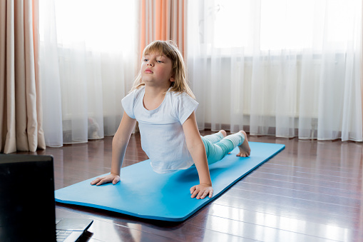 Little girl make home online laptop workout in flat.Baby yoga,fitness training.Distance school physical morning exercises on blue mat.Healthy activity,fun.Quarantine,coronavirus,covid-19.No equipment.