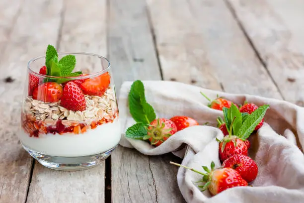 Yogurt with muesli and fresh strawberries for healthy breakfast or snack. Strawberry dessert parfait with yogurt and granola on a wooden table and linen kitchen-towel. Healthy and organic nutrition concept. Selective focus.