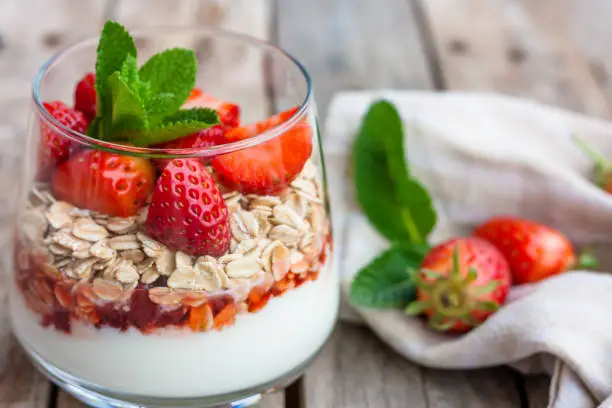 Yogurt with muesli and fresh strawberries for healthy breakfast or snack. Strawberry dessert parfait with yogurt and granola on a wooden table and linen kitchen-towel. Healthy and organic nutrition concept. Selective focus.