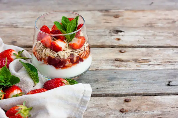 Yogurt with muesli and fresh strawberries for healthy breakfast or snack. Strawberry dessert parfait with yogurt and granola on a wooden table and linen kitchen-towel. Healthy and organic nutrition concept. Selective focus.