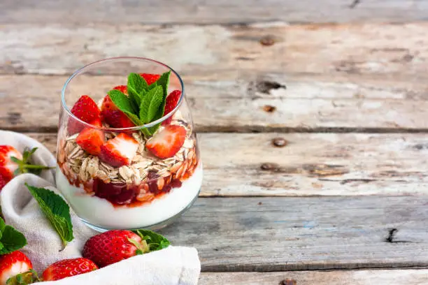 Yogurt with muesli and fresh strawberries for healthy breakfast or snack. Strawberry dessert parfait with yogurt and granola on a wooden table and linen kitchen-towel. Healthy and organic nutrition concept. Selective focus.