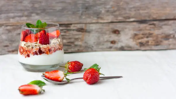 Yogurt with muesli and fresh strawberries for healthy breakfast or snack. Strawberry dessert parfait with yogurt and granola on a wooden table and linen kitchen-towel. Healthy and organic nutrition concept. Selective focus.