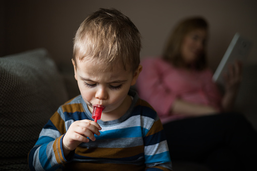 Young boy licking red lollipop while his mother sitting on the couch and staring at the tablet