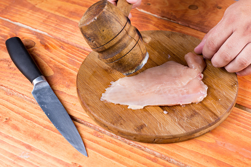 Chicken breasts fillet under wooden meat hammer on the board.